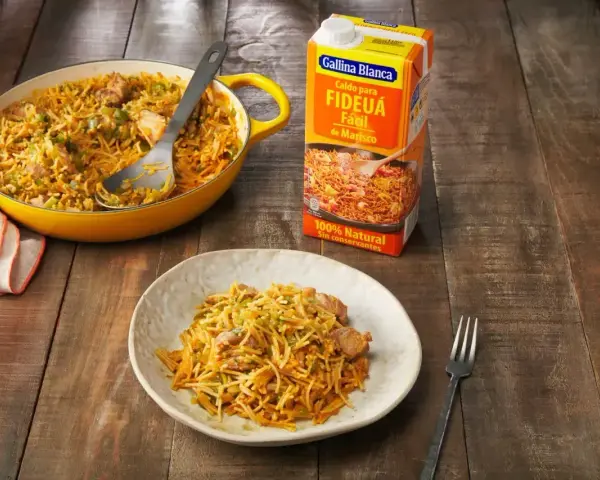 Un plato de fideos a la cazuela servido en una cazuela amarilla y en un plato blanco, junto a un cartón de caldo para fideuá.