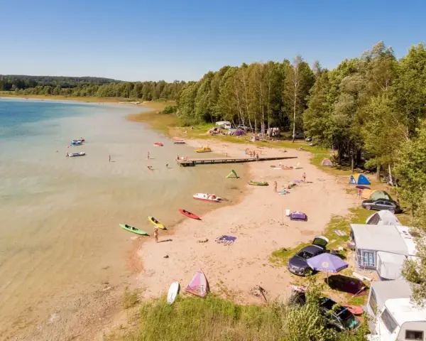 Letnie popołudnie nad jeziorem Świętajno. Ludzie kąpią się, pływają kajakami i odpoczywają na plaży wśród namiotów i kamperów.