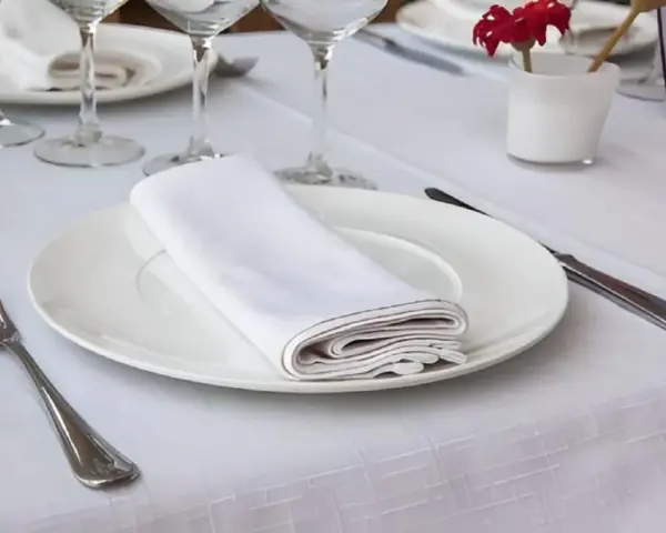 Une table dressée avec une assiette blanche, une serviette de table pliée, une fourchette et un couteau. La dimension serviette de table est parfaite pour cette présentation élégante.
