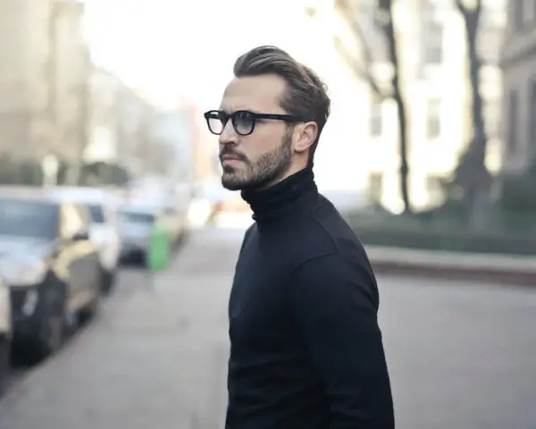 Elegancki mężczyzna w czarnym golfie i okularach, zamyślony, jakby planował zagrywki zakochanego faceta.