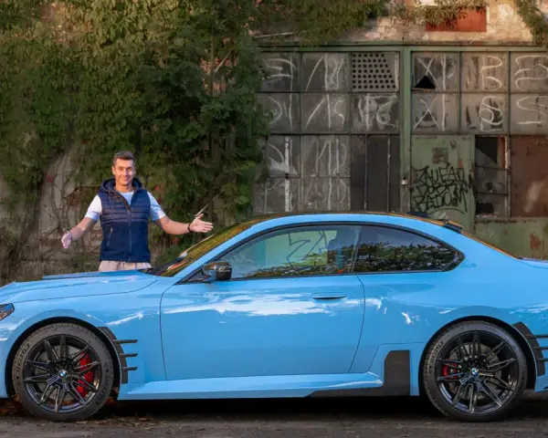 Mężczyzna prezentuje błękitne BMW M2 G87 przed zniszczonym budynkiem.