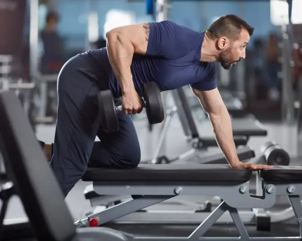 Man utför enarms hantelrodd på ett gym. Han stöttar sig mot en bänk med ena handen och drar en hantel mot bröstet med den andra.