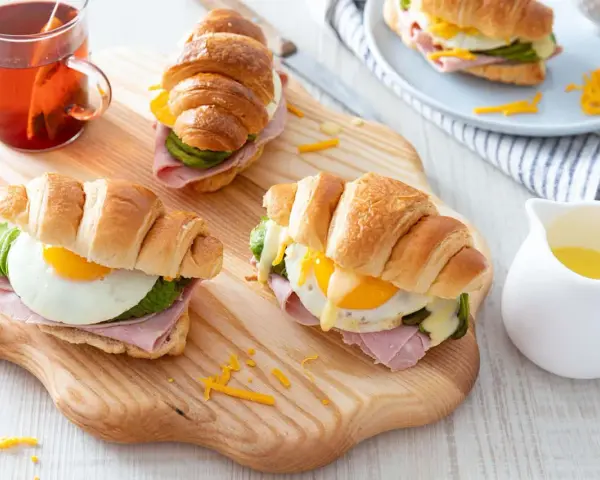 Trois croissants au jambon, œuf au plat, avocat et fromage râpé, servis sur une planche en bois avec une tasse de thé.