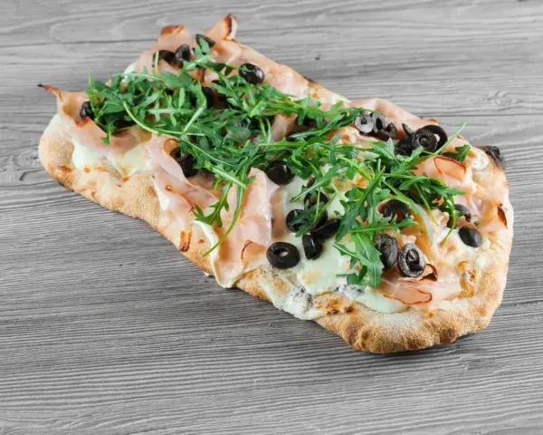 Pyszne ciasto na pizzę z szynką, mozzarellą, rukolą i czarnymi oliwkami, ułożone na drewnianym blacie.