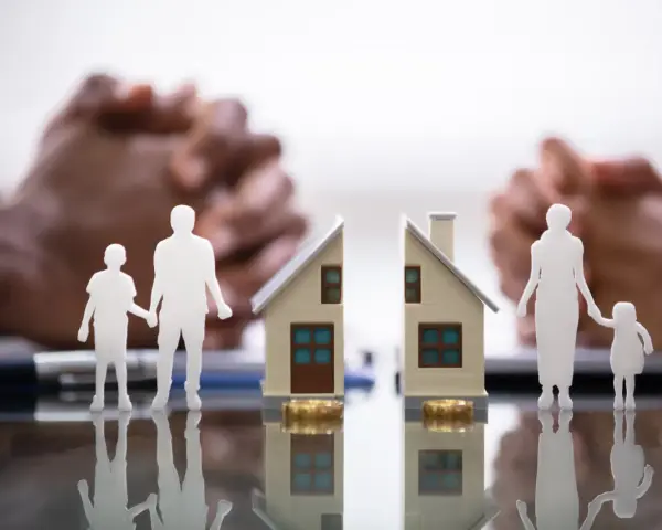 Figurines de famille séparées par une maison coupée en deux, symbolisant une convention d'indivision divorce. Mains floues en arrière-plan.