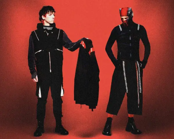 Dwóch artystów w czarnych strojach, przypominających styl z okładek płyt twenty one pilots, pozuje na tle czerwonym.