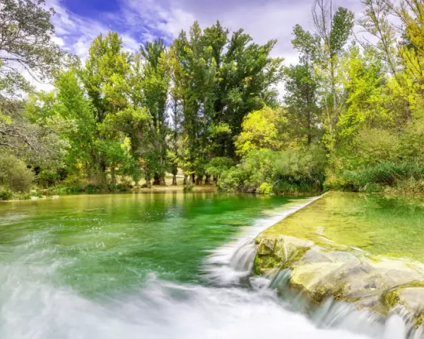 La poza de doña urraca, un remanso de agua verde esmeralda con una pequeña cascada, rodeada de frondosa vegetación y árboles altos bajo un cielo azul.