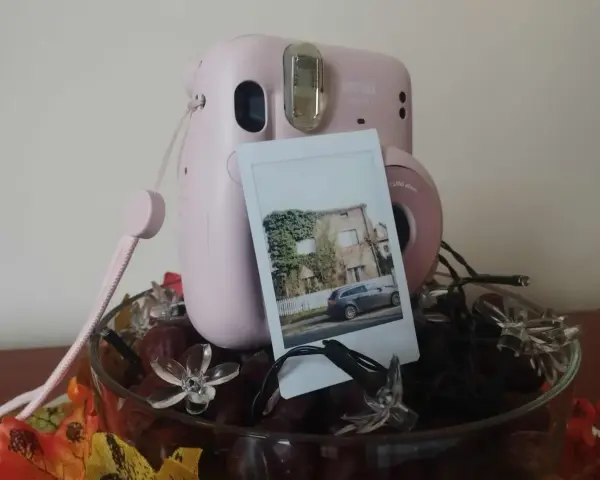 Różowy aparat Instax z wydrukowanym zdjęciem domu i samochodu. Idealny pomysł, jak zrobić prezent ze zdjęć, ozdobiony jesiennymi liśćmi i lampkami.
