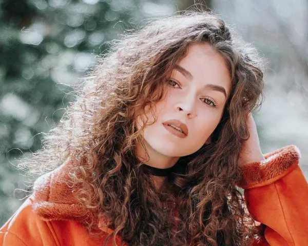 Jeune femme aux cheveux bouclés, portant une veste orange. La retouche photo netteté en ligne sublime son regard intense.