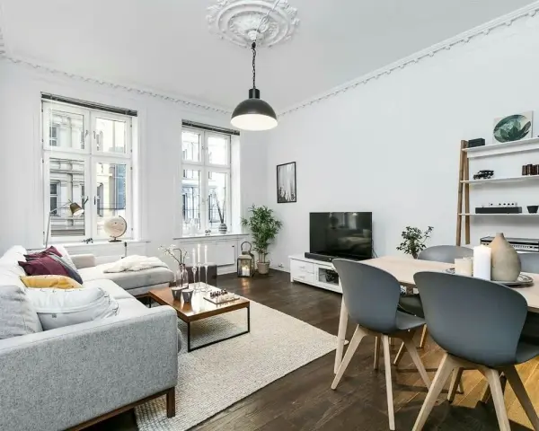 Elegante salón con sofá gris, mesa de centro y comedor. El **suelo oscuro** de madera contrasta con las paredes blancas y la luz natural.