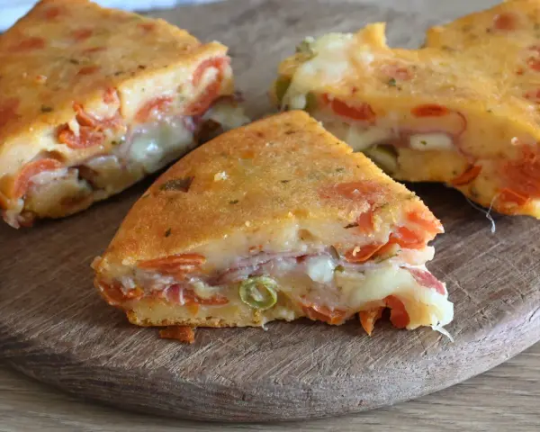 Fette di torta salata senza uova, ricca di pomodoro, formaggio filante e prosciutto, su un tagliere di legno.