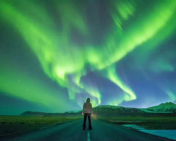 Człowiek stoi na drodze pod rozgwieżdżonym niebem, gdzie tańczy zorza polarna. Islandia, październik, pogoda idealna na takie widowisko.