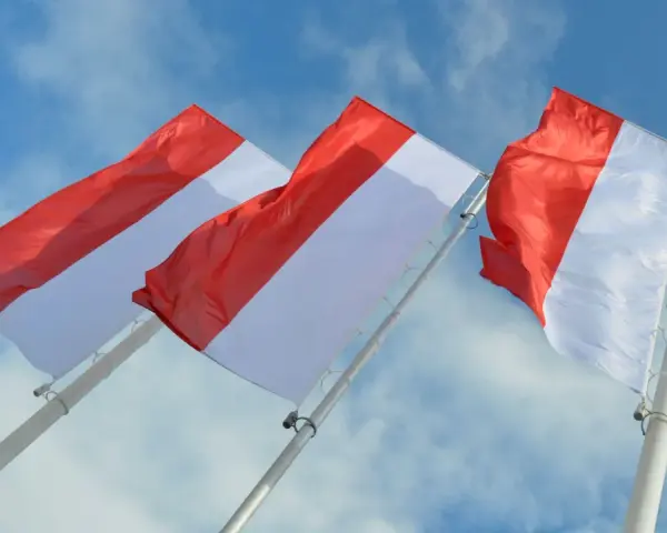 Trzy powiewające na wietrze flagi, każda z czerwono-białą szarfą, na tle błękitnego nieba z chmurami.