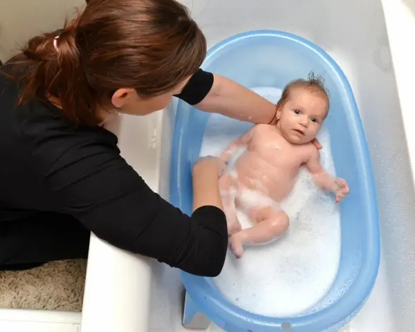 Mama delikatnie myje noworodka w niebieskiej wanience. Maluch pluska się w pianie, a mama uśmiecha się do niego.