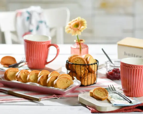 Leckeres Zwieback-Rezept: goldbraune Scheiben auf rosa Platte, dazu Marmelade, Butterdose und rote Tassen.