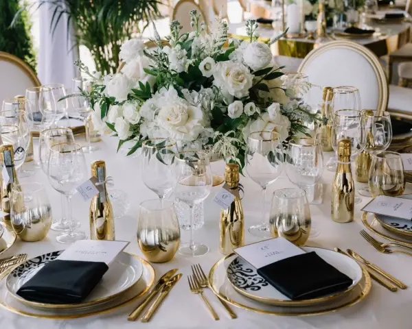 Elegancka kompozycja na stół weselny: białe kwiaty, złote buteleczki i szkło.