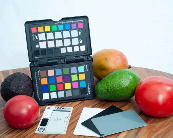 Na drewnianym blacie leżą owoce: awokado, mango, pomidory. Obok znajduje się otwarty colorchecker z kolorowymi polami i szara karta do kalibracji balansu bieli.