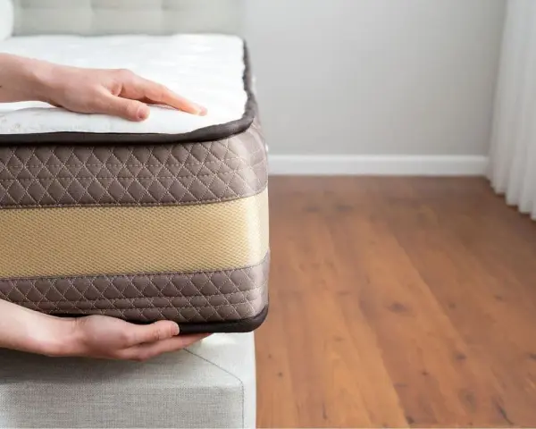 Mains posées sur un matelas, montrant sa bonne épaisseur. Le tissu matelassé marron et la bande beige suggèrent un confort optimal.