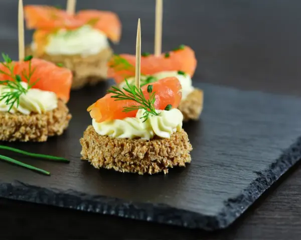 Eleganckie przystawki na komunię: krążki ciemnego pieczywa z kremowym serkiem, łososiem i koperkiem.