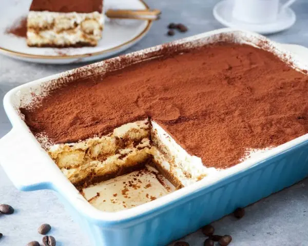 Ein köstliches Tiramisu rezept ohne alkohol, bestäubt mit Kakao, in einer blauen Auflaufform. Ein Stück ist bereits entnommen.