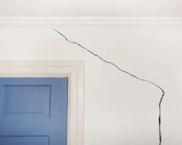 Grosse fissure sur mur intérieur blanc, près d'une porte bleue. Il faut réparer cette fissure pour un intérieur impeccable.