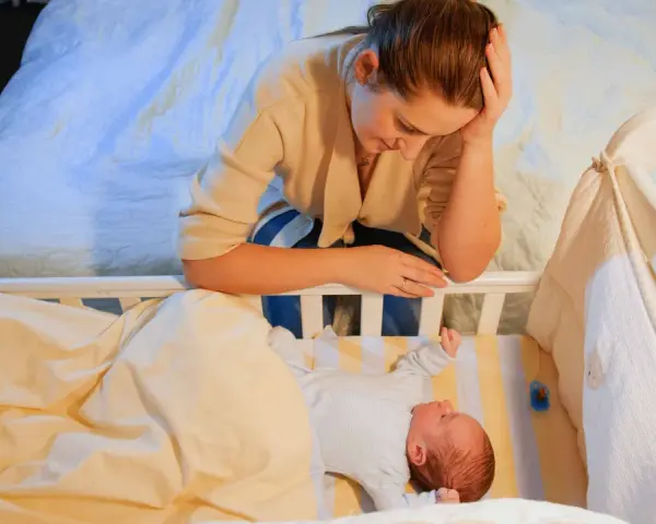 Zmęczona mama pochyla się nad łóżeczkiem, zastanawiając się, kiedy nastąpi regres snu. Niemowlę śpi spokojnie.