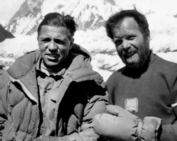 Zwei Bergsteiger, die die K2 Erstbesteigung schafften, lächeln in die Kamera. Im Hintergrund sind schneebedeckte Berge zu sehen.