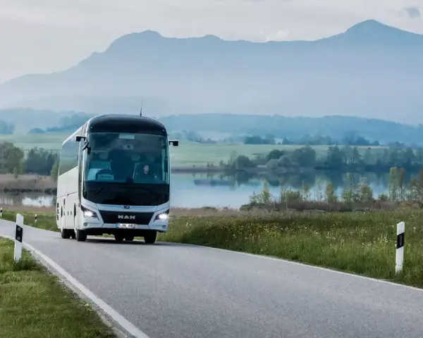 Biały **bus** marki **MAN** jedzie krętą drogą nad jeziorem, w tle góry.