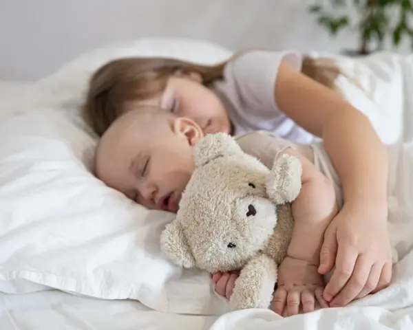 Dzieci śpią wtulone w misia. Starsza siostra czuwa nad maluszkiem, pokazując, ile śpi dwulatek i jak ważny jest sen dla rozwoju.
