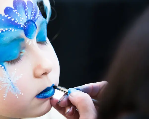 Ein Kind wird mit blauem Lidschatten und Schneeflocken bemalt, um wie Elsa auszusehen. Das Schminken ist leicht und zaubert ein magisches Wintergesicht.