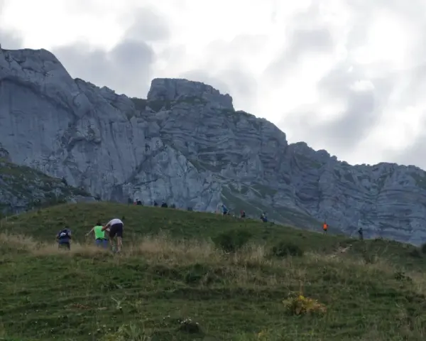 Randonneurs sur une pente herbeuse face à une falaise imposante. Le chemin sinueux et la hauteur suggèrent que la tournette est dangereuse.