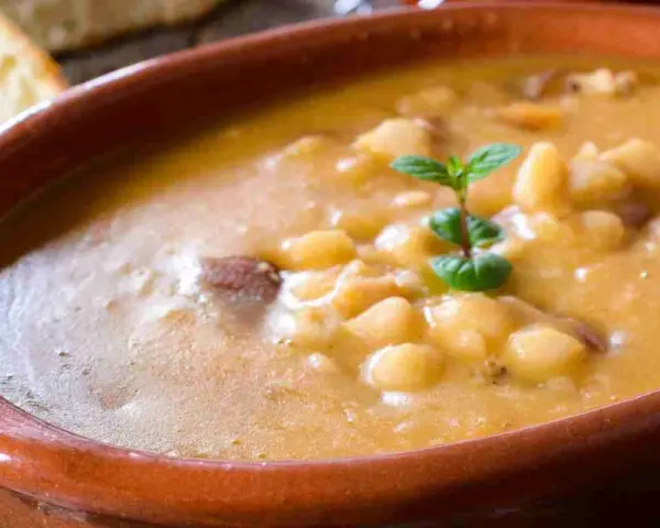 Una ciotola di terracotta piena di zuppa di fagioli cremosa, con pezzetti di carne e una fogliolina di menta. Perfetta per una cena confortante.