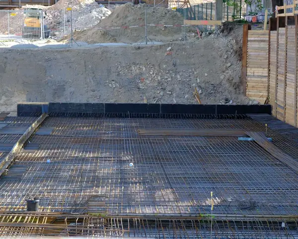Armatura per fondazione a platea sezione. La rete metallica è pronta per il getto di calcestruzzo, con terra e muri di contenimento sullo sfondo.