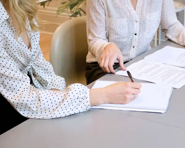 Two women in polka-dot shirts review documents. One takes notes, implying they are discussing who can see committee meeting minutes.