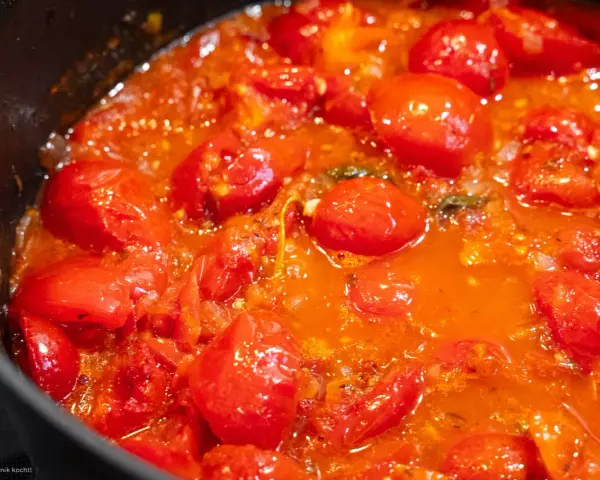 Ein köstliches, blubberndes Tomaten Sugo in einer schwarzen Pfanne, voller saftiger Kirschtomaten und aromatischer Kräuter.