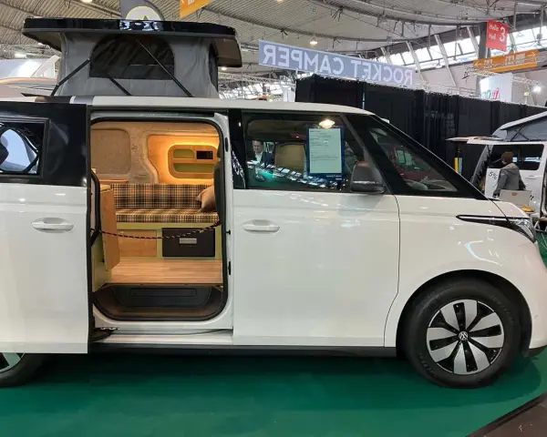 Vit ID Buzz camper van med uppfällt tak och öppen skjutdörr som visar en mysig interiör med rutig soffa och trädetaljer.
