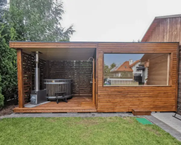 Drewniany domek z sauną i beczką do kąpieli, z piecem zewnętrznym i prysznicem. Duże okno wychodzi na ogród.