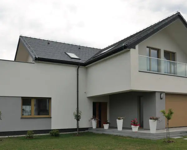 Nowoczesny dom z kremową elewacją, szarym cokołem, dużym oknem i balkonem ze szklaną balustradą. Przed wejściem białe donice z kwiatami.
