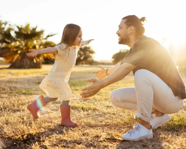 Ein kleines Mädchen rennt mit offenen Armen auf ihren lachenden Vater zu. Die warme Abendsonne beleuchtet die Szene und unterstreicht die liebevolle vater tochter beziehung.