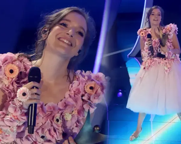 Zuzanna Irena Jurczak w kwiecistej sukni na scenie, trzyma mikrofon i uśmiecha się do publiczności.