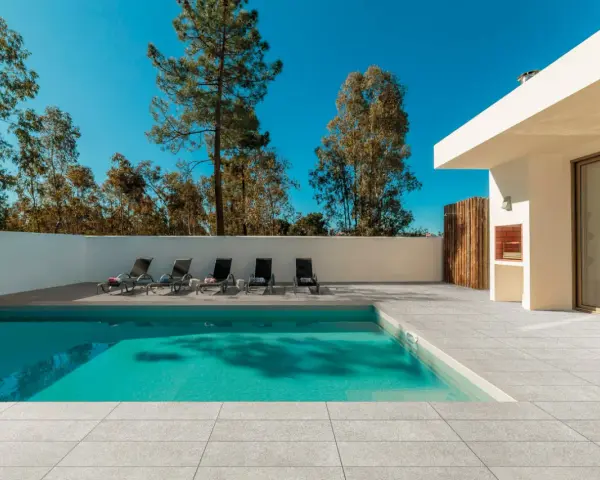 Une piscine moderne avec un sol en carrelage gris clair, des chaises longues et un barbecue extérieur sous un ciel bleu éclatant.