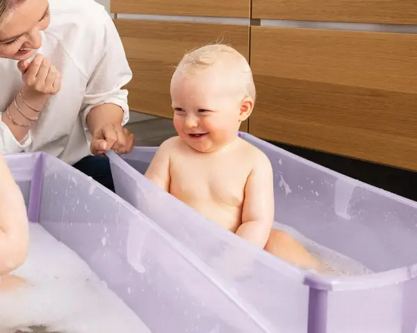 Uśmiechnięty bobas w fioletowym krzesełku do kąpieli, mama patrzy z czułością.