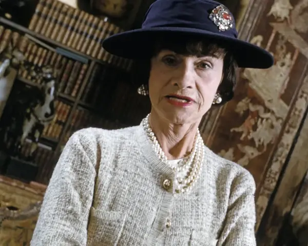 Iconica stilista Coco Chanel, con cappello blu e perle, in un ambiente elegante con libri e decorazioni.
