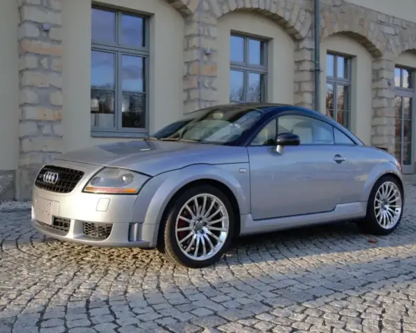 Silberner Audi TT 2005 mit schwarzem Dach parkt auf Kopfsteinpflaster vor einem Gebäude mit Steinbögen.