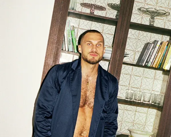 Hombre con pelo rapado y barba, viste una chaqueta abierta sobre el pecho. Detrás, una estantería con libros y vasos.