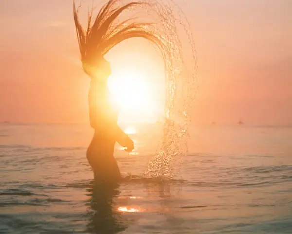 Silhouette d'une femme jetant ses cheveux dans l'eau au coucher du soleil, avec un effet de lens flare photoshop.