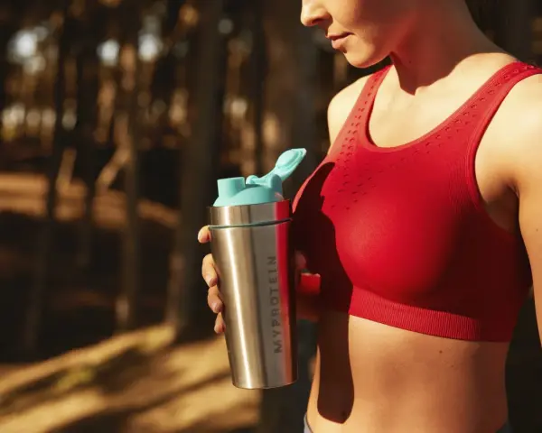 En kvinna i röd sporttopp håller en proteinshaker. Frågan om hur mycket protein kan kroppen ta upp är relevant för hennes träning.