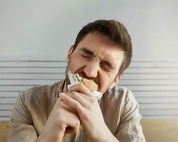 Uomo con espressione sofferente mangia un panino, forse per alleviare lo stress, ma teme di ingrassare.