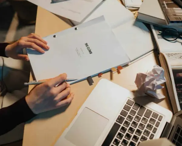 Manos escribiendo un guion cortometraje, rodeado de libros, gafas y un portátil.