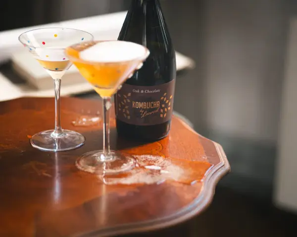 Dwa kieliszki koktajlowe z pianką i butelka kombuchy na drewnianym stole. Czy kombucha ma alkohol? Ta butelka sugeruje, że może mieć.